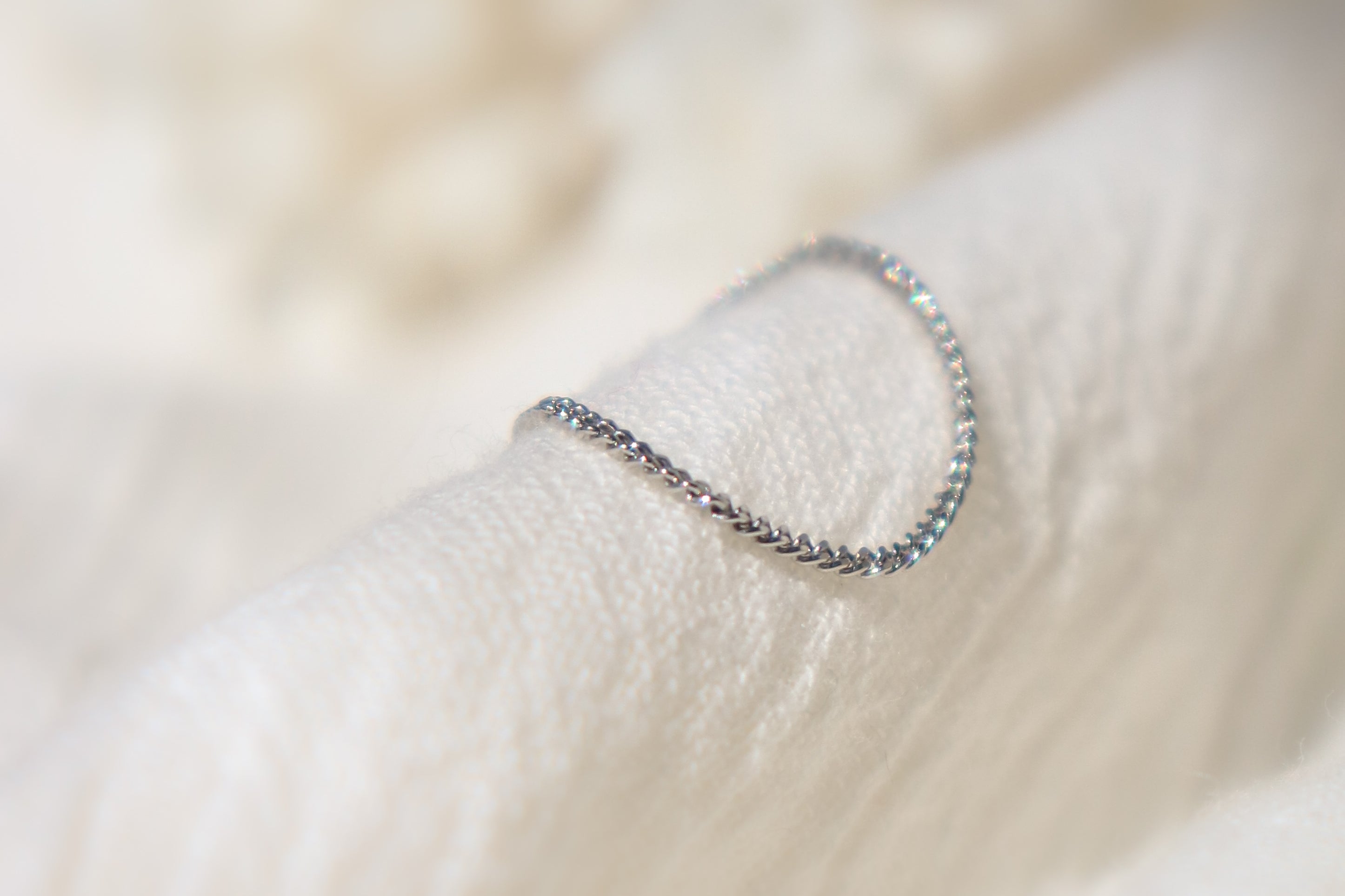 Delicate platinum chain link ring with small glints of light reflecting off the surface on a soft beige fabric background