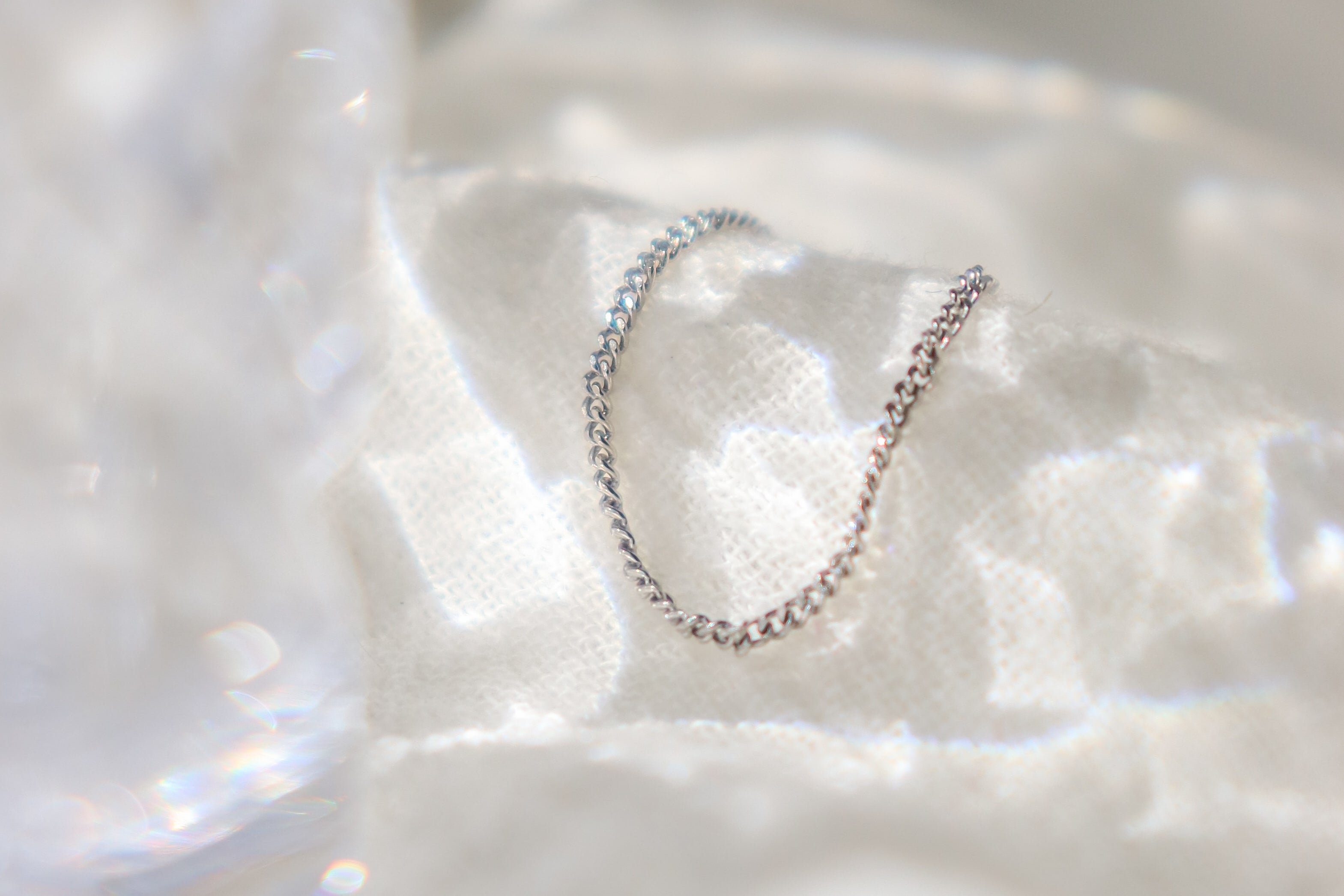 Platinum chain link finger ring for women on a textured white surface with water like shadows reflecting off the background and a crystal glass blurred out off to the side.