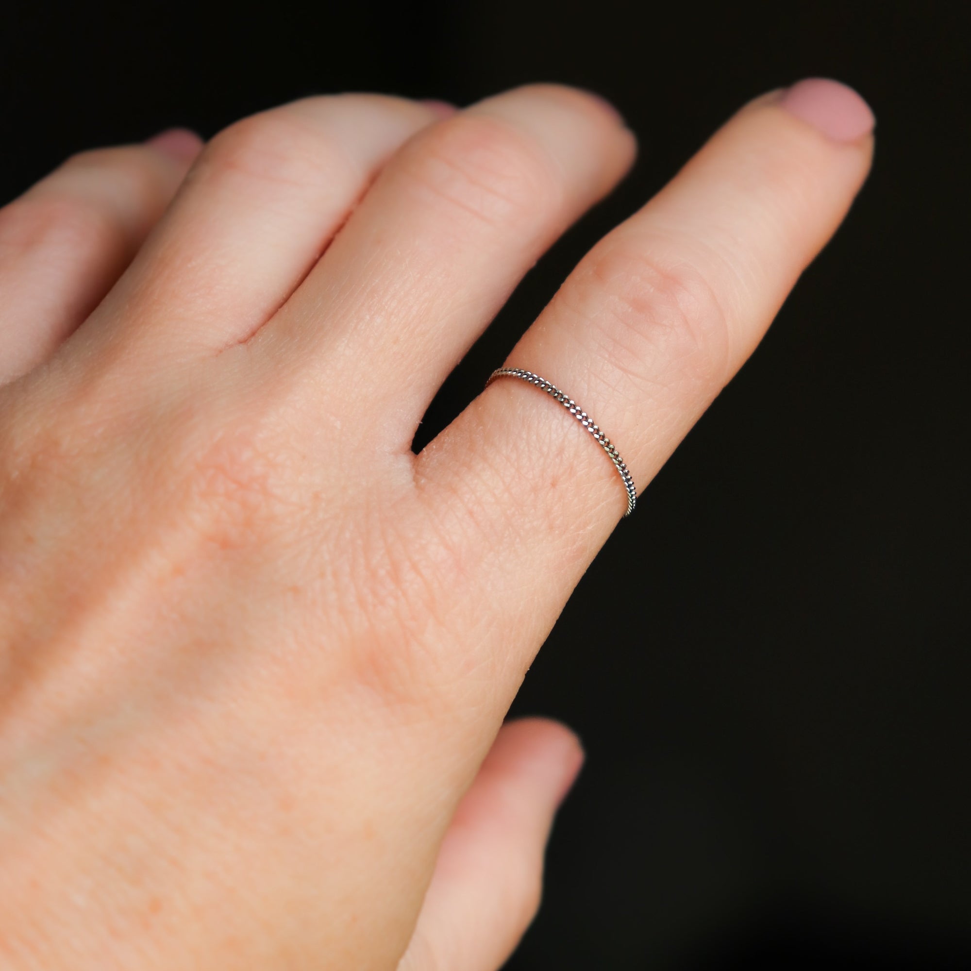 Close up of a woman's hand wearing a petite platinum chain ring on a dark background.