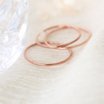 Three dainty, smooth, rose gold stackable rings are laying on top of each other on a light fabric background with blurred crystal glass in the background.