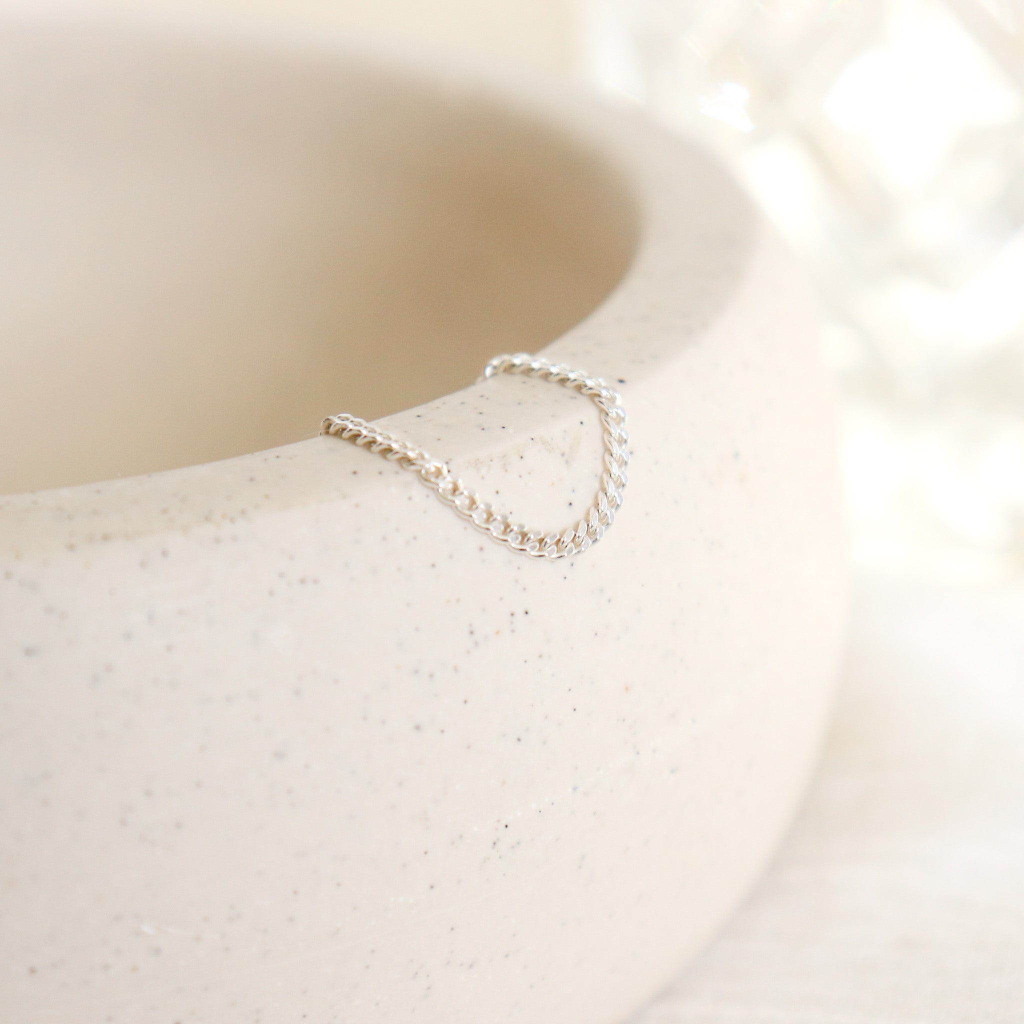 Women's dainty silver chain link ring draped over the side of a pottery dish with a soft beige background