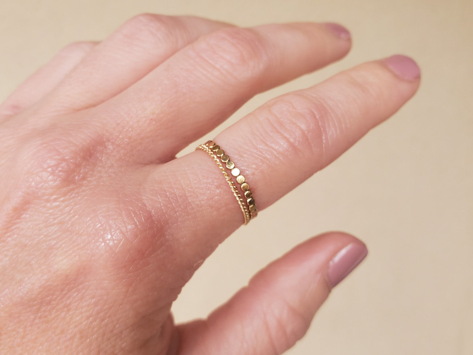Hand wearing a gold beaded ring and a gold chain ring on a beige background