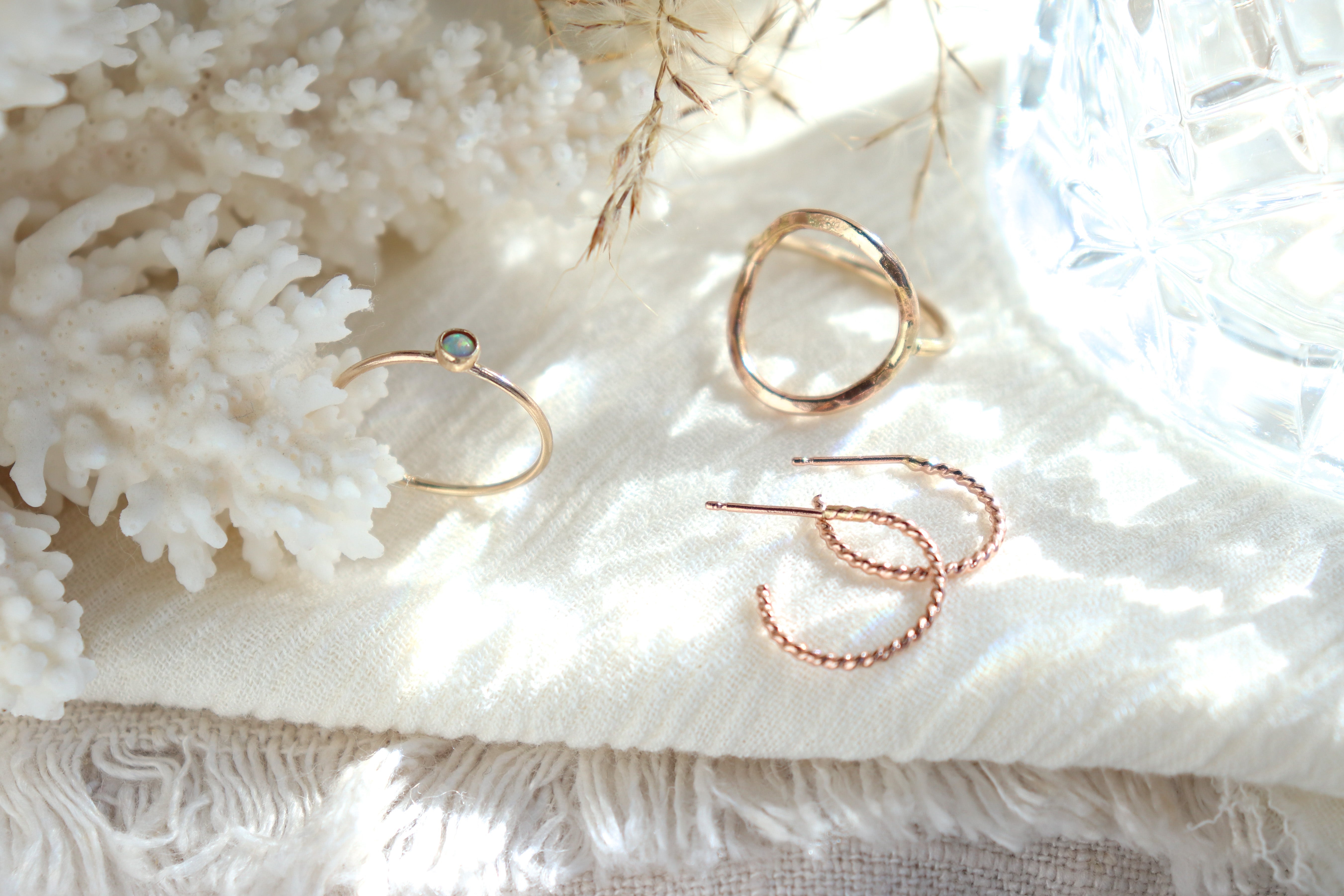 Several pieces of dainty, minimalist jewelry including a gold opal ring, round gold hammered statement ring and a pair of small rose gold huggie hoop earrings are displayed on a light background consisting of a linen fabric hi lighted by scattered light from a crystal glass and soft coral and dried floral in the background.