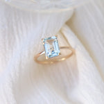 Gold prong set ring featuring a 6x8 mm emerald cut natural aquamarine gemstone on a light fabric background.