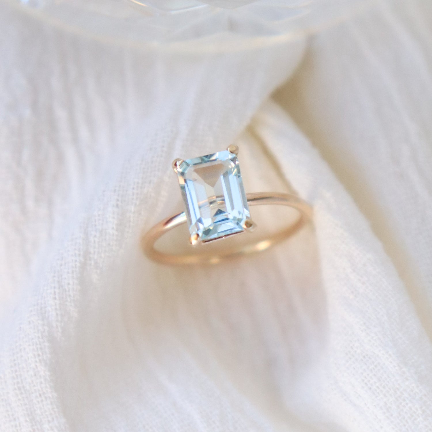 Gold prong set ring featuring a 6x8 mm emerald cut natural aquamarine gemstone on a light fabric background.