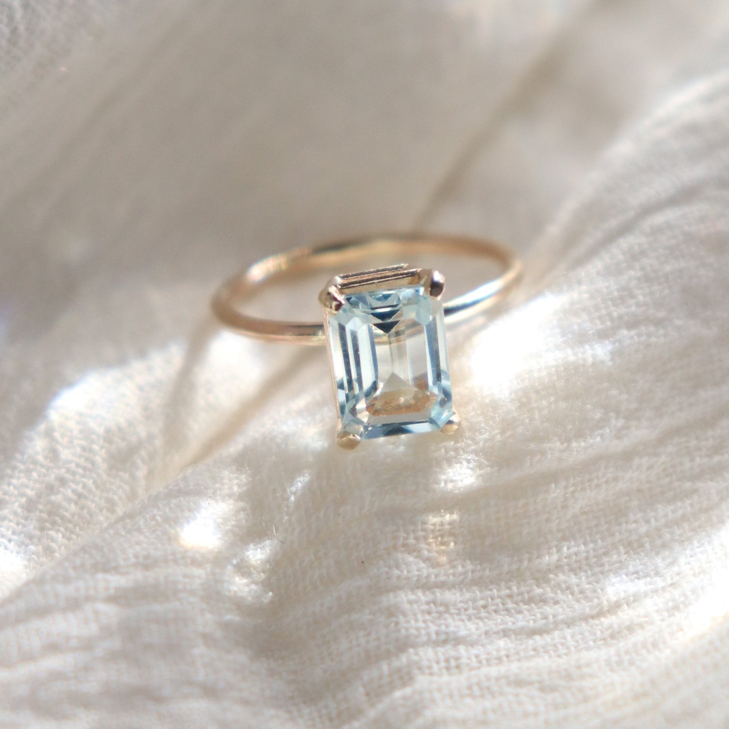Gold prong set emerald cut large aquamarine solitaire ring with a rectangular blue gemstone on a white fabric background