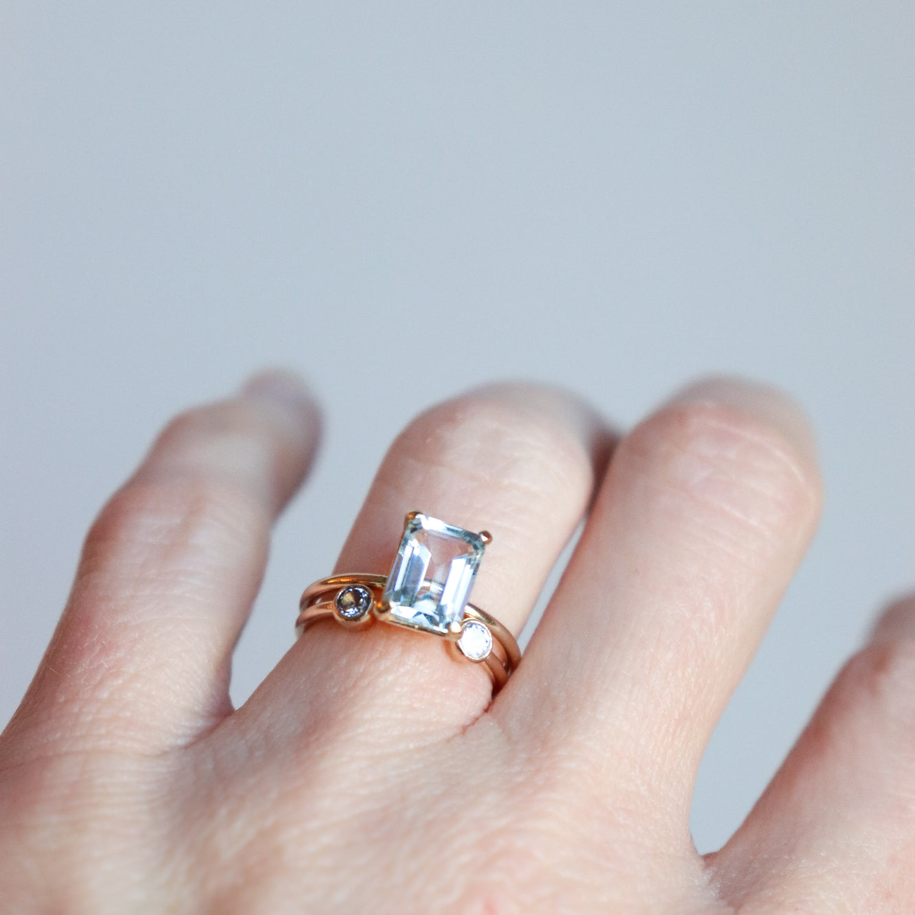Woman's hand wearing a rose gold ring with a large, natural aquamarine emerald-cut gemstone on a light gray background. The featured ring is paired with a smaller dual luminary birthstone ring by Meadowbelle.