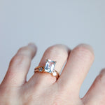 Woman's hand wearing a rose gold ring with a large, natural aquamarine emerald-cut gemstone on a light gray background. The featured ring is paired with a smaller dual luminary birthstone ring by Meadowbelle.