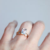 Woman's hand wearing a rose gold ring with a large, natural aquamarine emerald-cut gemstone on a light gray background. The featured ring is paired with a smaller dual luminary birthstone ring by Meadowbelle.