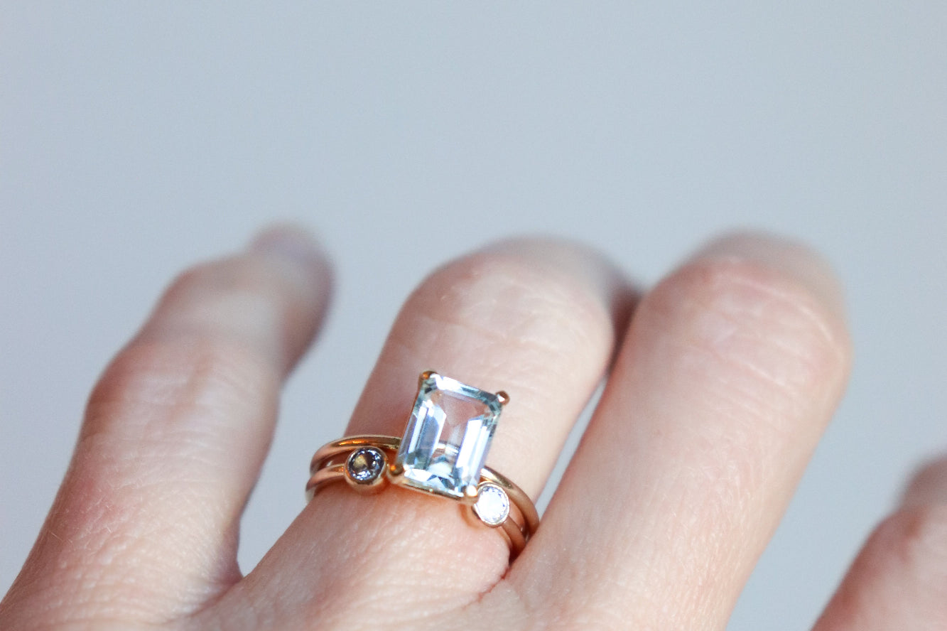 Woman's hand wearing a rose gold ring with a large, natural aquamarine emerald-cut gemstone on a light gray background. The featured ring is paired with a smaller dual luminary birthstone ring by Meadowbelle.