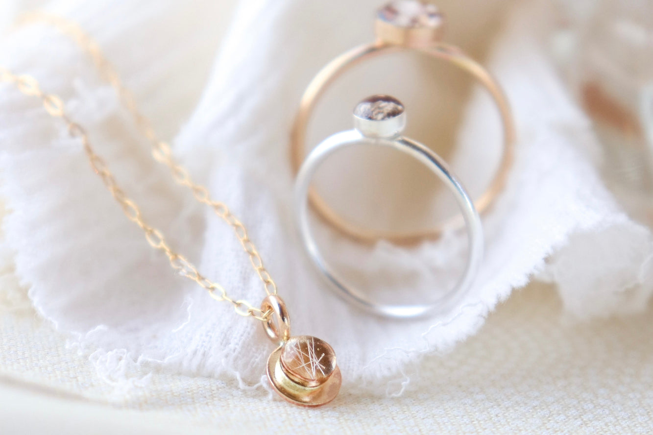 Gold memorial necklace and two memorial rings in silver and gold balanced upright on a light creamy and white fabric background.