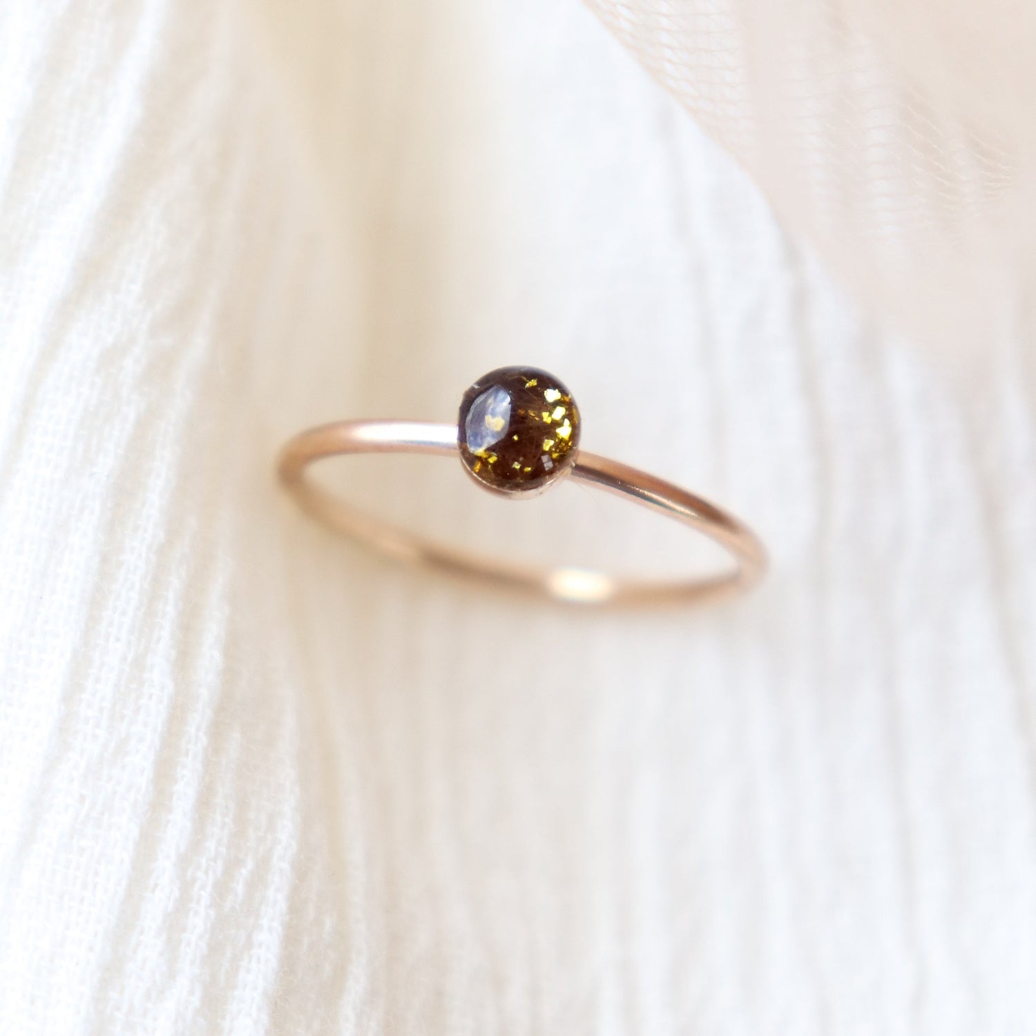 Small gold memorial ring with a dark chocolate colored resin ashes cabochon with gold flecks. The ring is balanced on a light background. By Meadowbelle Jewelry.