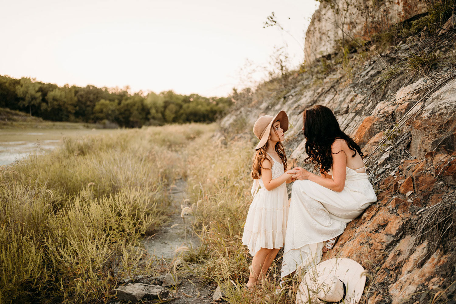Mother and daughter wearing light, flown dresses holding hands while looking at each other. They are wearing dainty, minimalist jewelry. The backdrop is a field with rocky hillside and tall grasses.