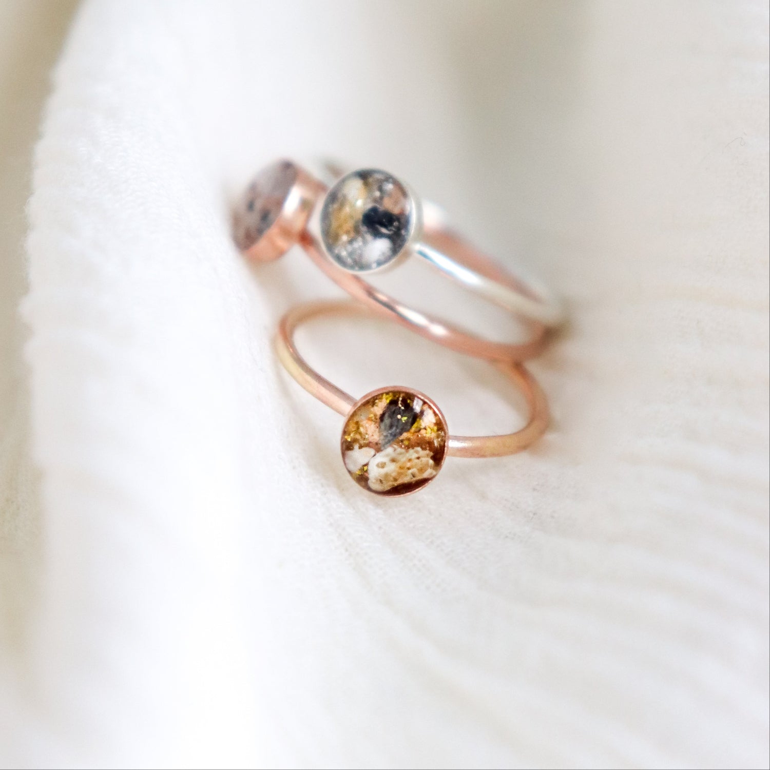 a stack of three mixed metal cremation ashes keepsake rings piled on a light fabric background. All the rings feature visible ashes in natural earth tones.