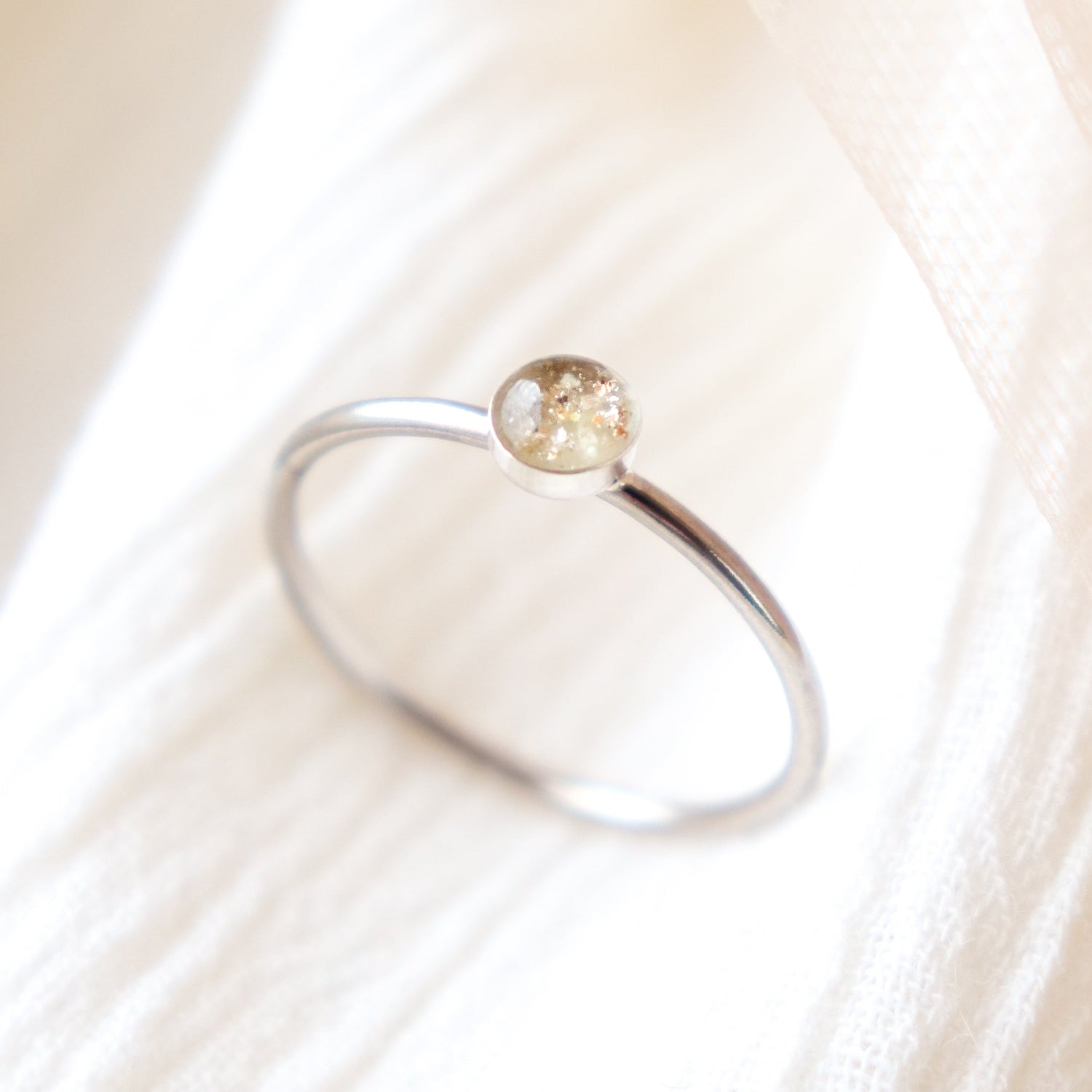 Platinum memorial ring sitting balanced upright with light colored ashes in a 4 mm clear resin setting. Featured on a soft beige fabric background.