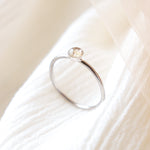 Platinum memorial ring with light colored ashes in a 4 mm clear resin setting. Featured sitting upright to see the size of the bezel setting on a soft beige fabric background.