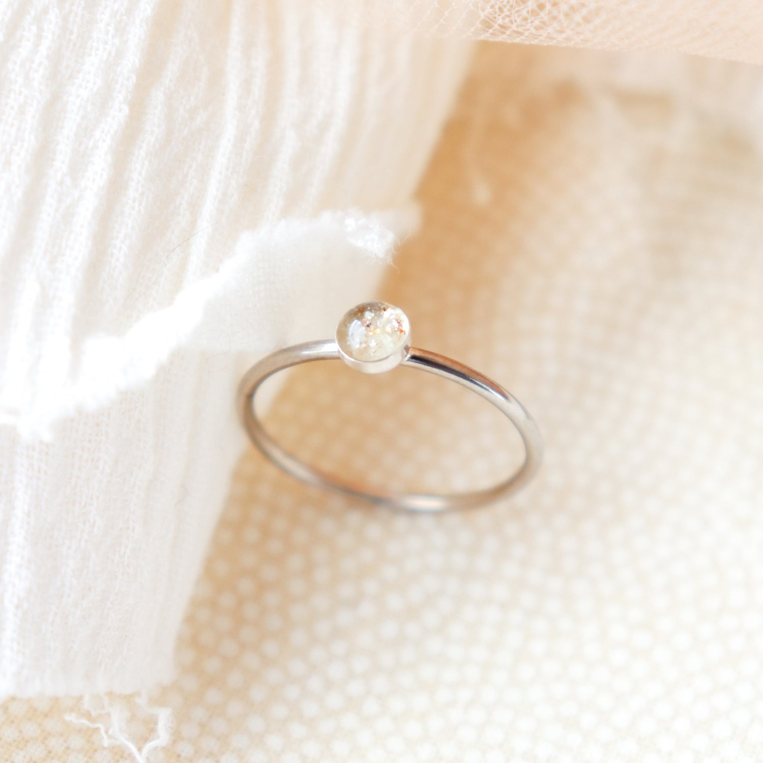 Platinum memorial ring with light colored ashes in a 4 mm clear resin setting. Featured on a soft beige fabric background.