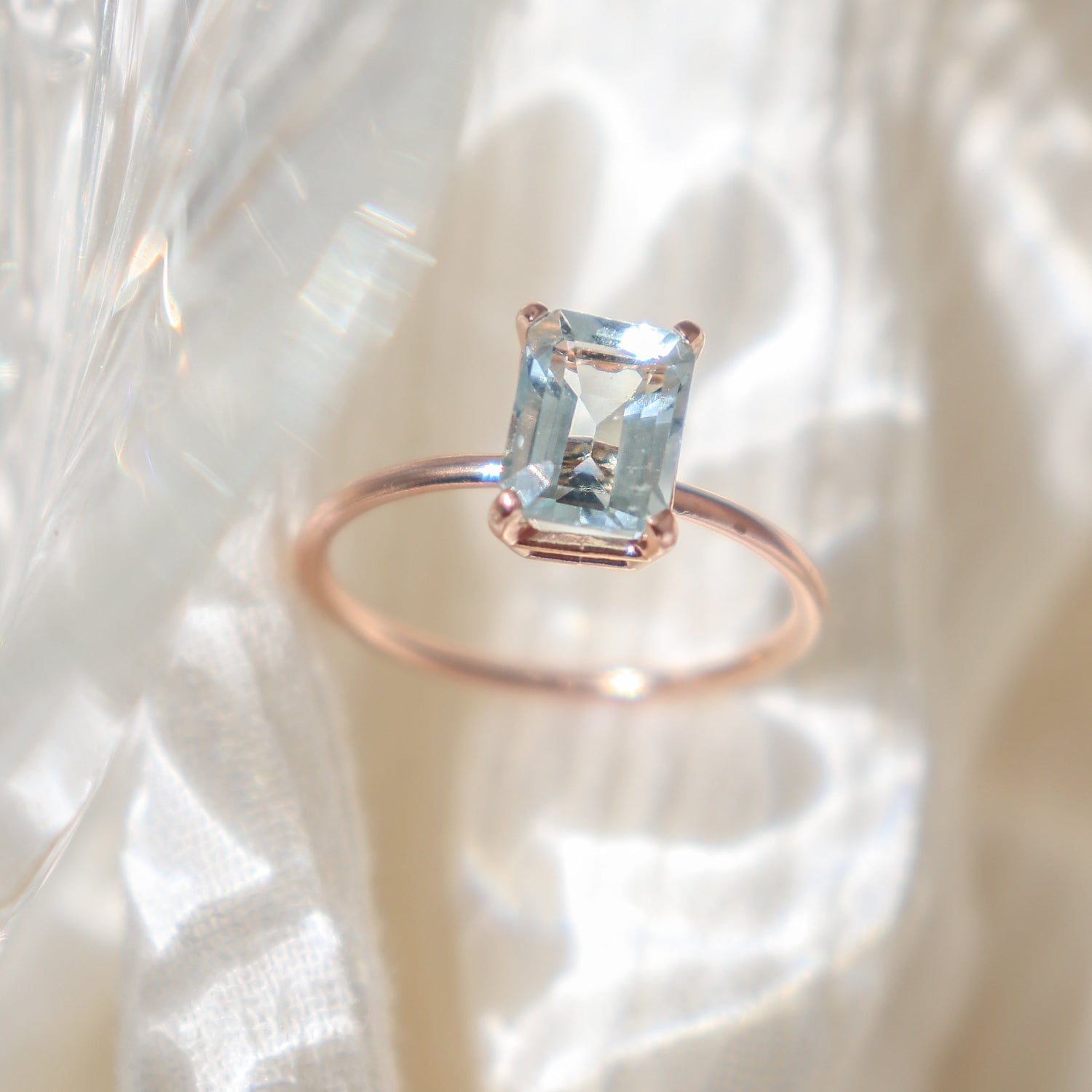 Large rose gold prong set emerald cut blue aquamarine solitaire ring in mottled shadowy light on a white fabric background.