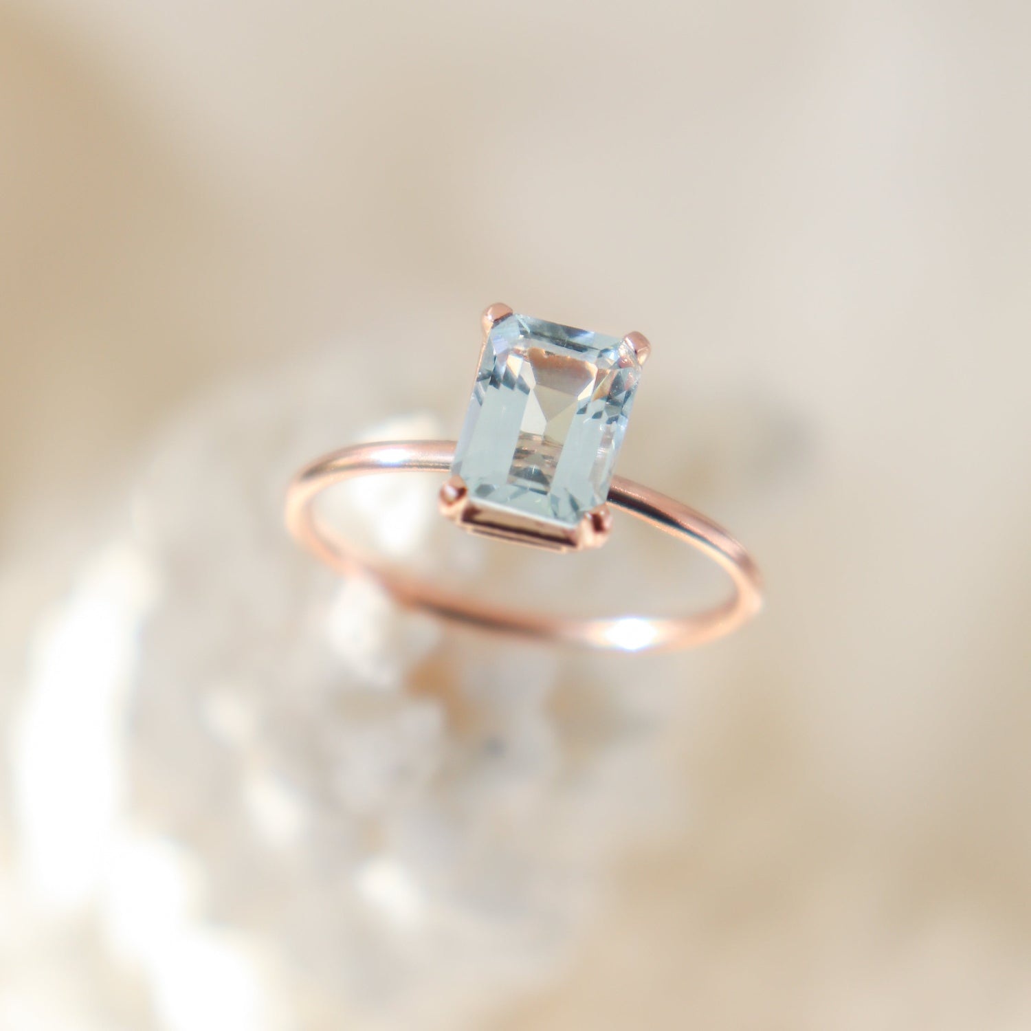 Large rose gold prong set emerald cut blue aquamarine solitaire ring balanced on a light, creamy, blurred background.