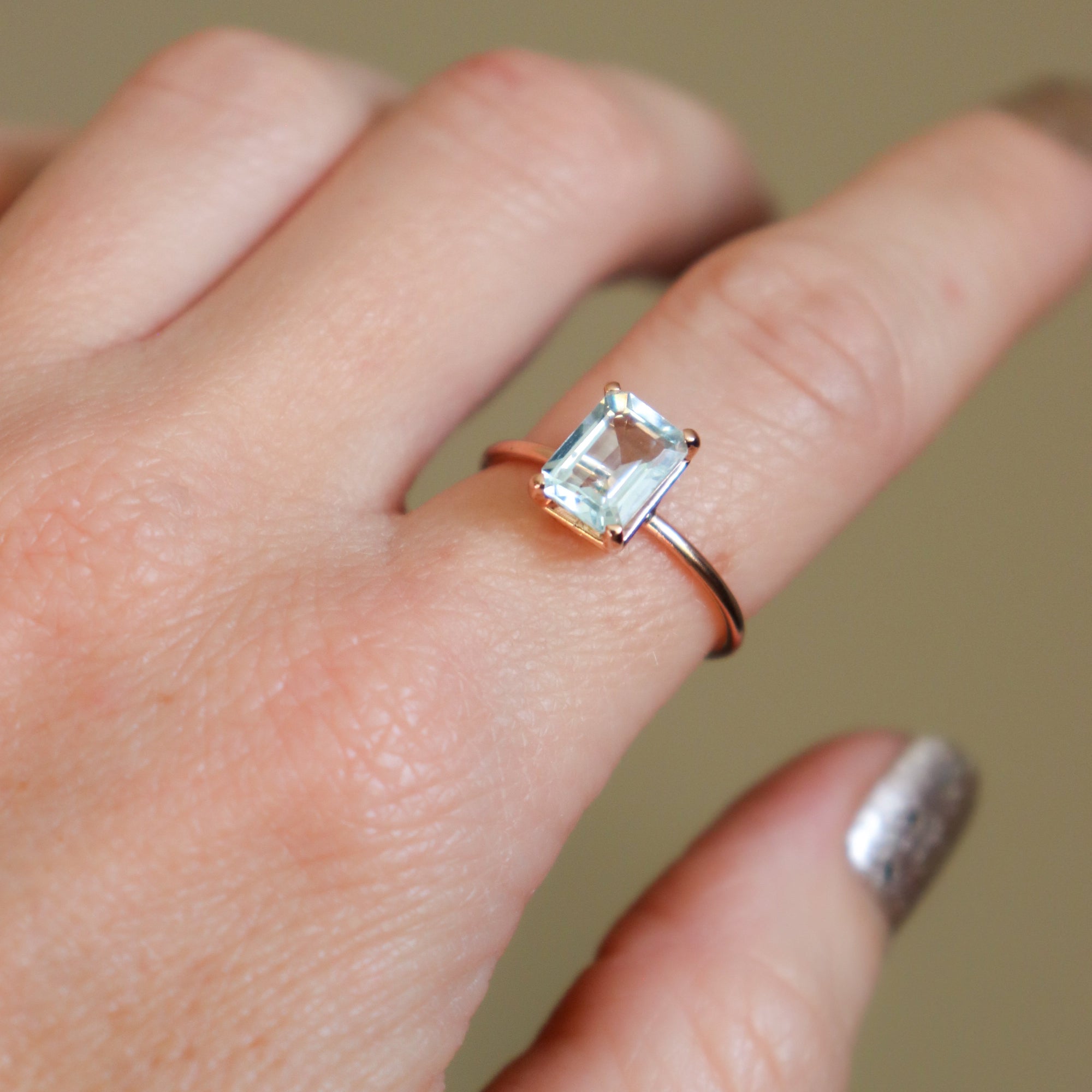 Hand wearing a large rose gold women's solitaire ring with a rectangular emerald cut natural aquamarine gemstone on a neutral background.