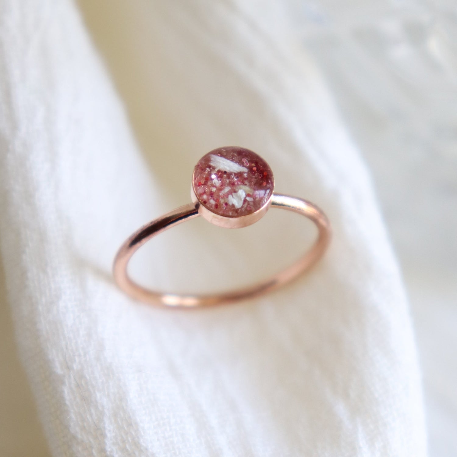 Close up of a solid 14k rose gold ashes cremation keepsake ring on a light fabric background. The coloring in the setting features ashes with light pink.