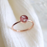 Close up of a solid 14k rose gold ashes cremation keepsake ring on a light fabric background. The coloring in the setting features ashes with light pink.