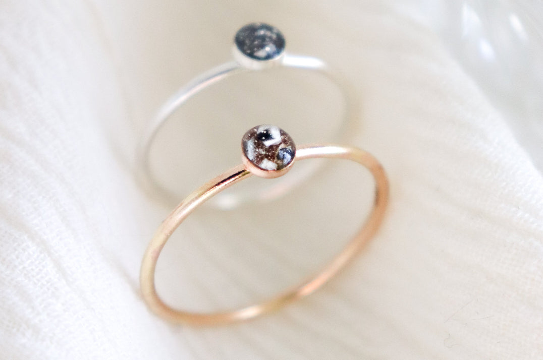 Two hand made, minimalist memorial rings on a light background. One ring is silver and the other ring is gold fill. Small, round resin cabochons hold ashes inclusions with custom color added.
