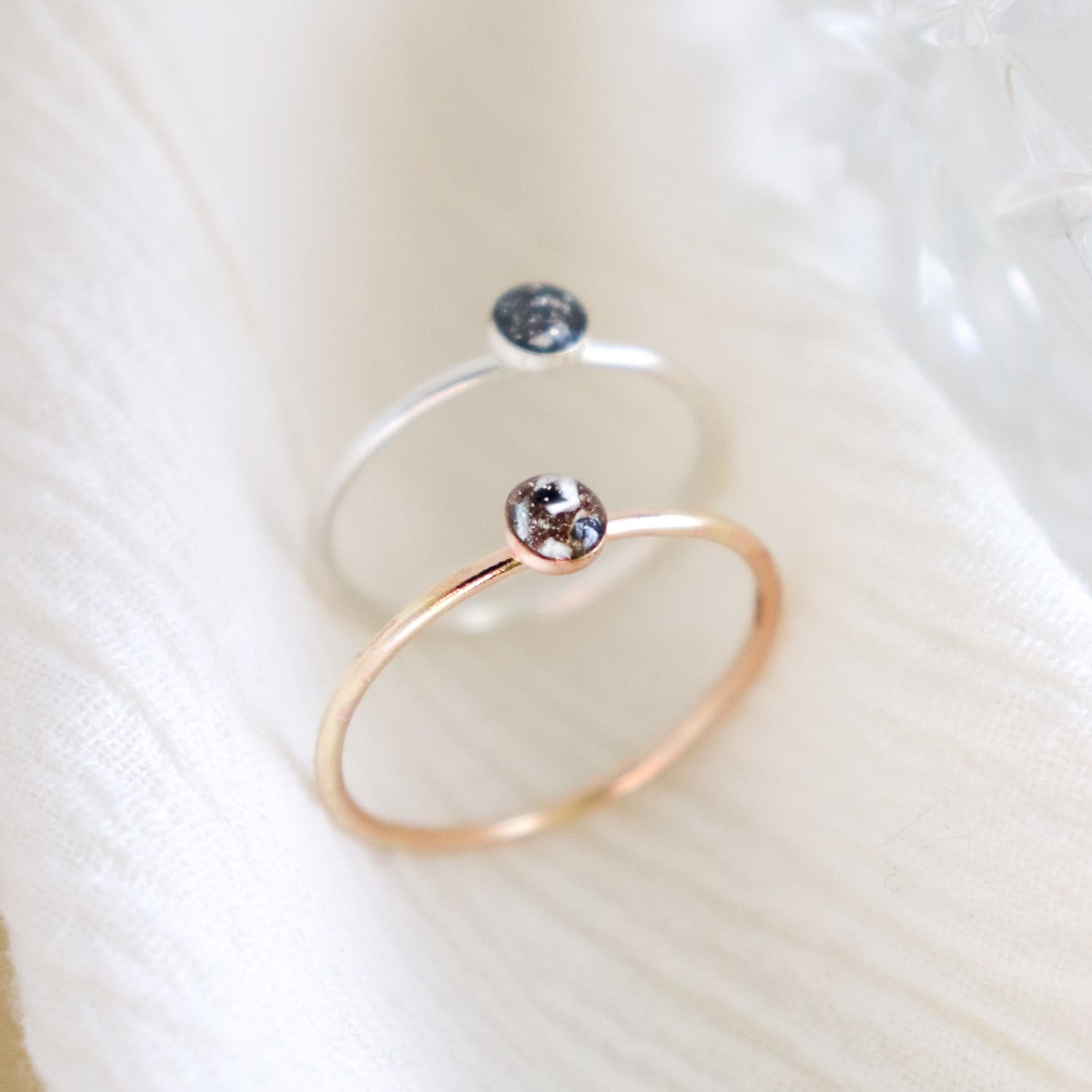 Two hand made, minimalist memorial rings on a light background. One ring is silver and the other ring is gold fill. Small, round resin cabochons hold cremated ashes with custom color inclusions.
