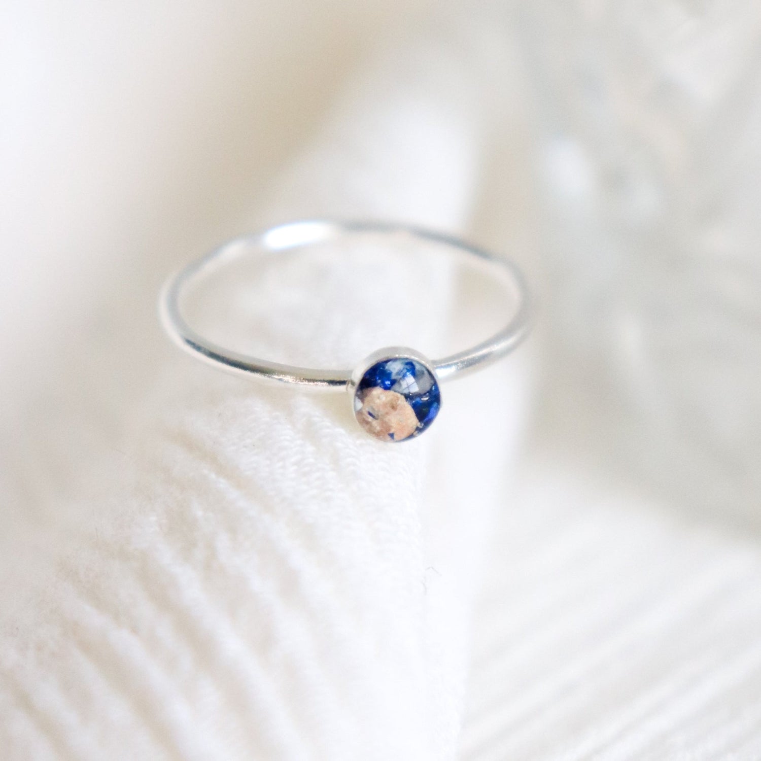 Small, minimalist memorial ring in sterling silver holds visible ashes in a petite round setting with Sapphire blue color included. The ring is on a light background.
