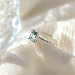 Rectangle shaped natural moss agate gemstone ring in a solid sterling silver setting with a smooth, round band. The ring is poised on a soft fabric background with moody lighting.