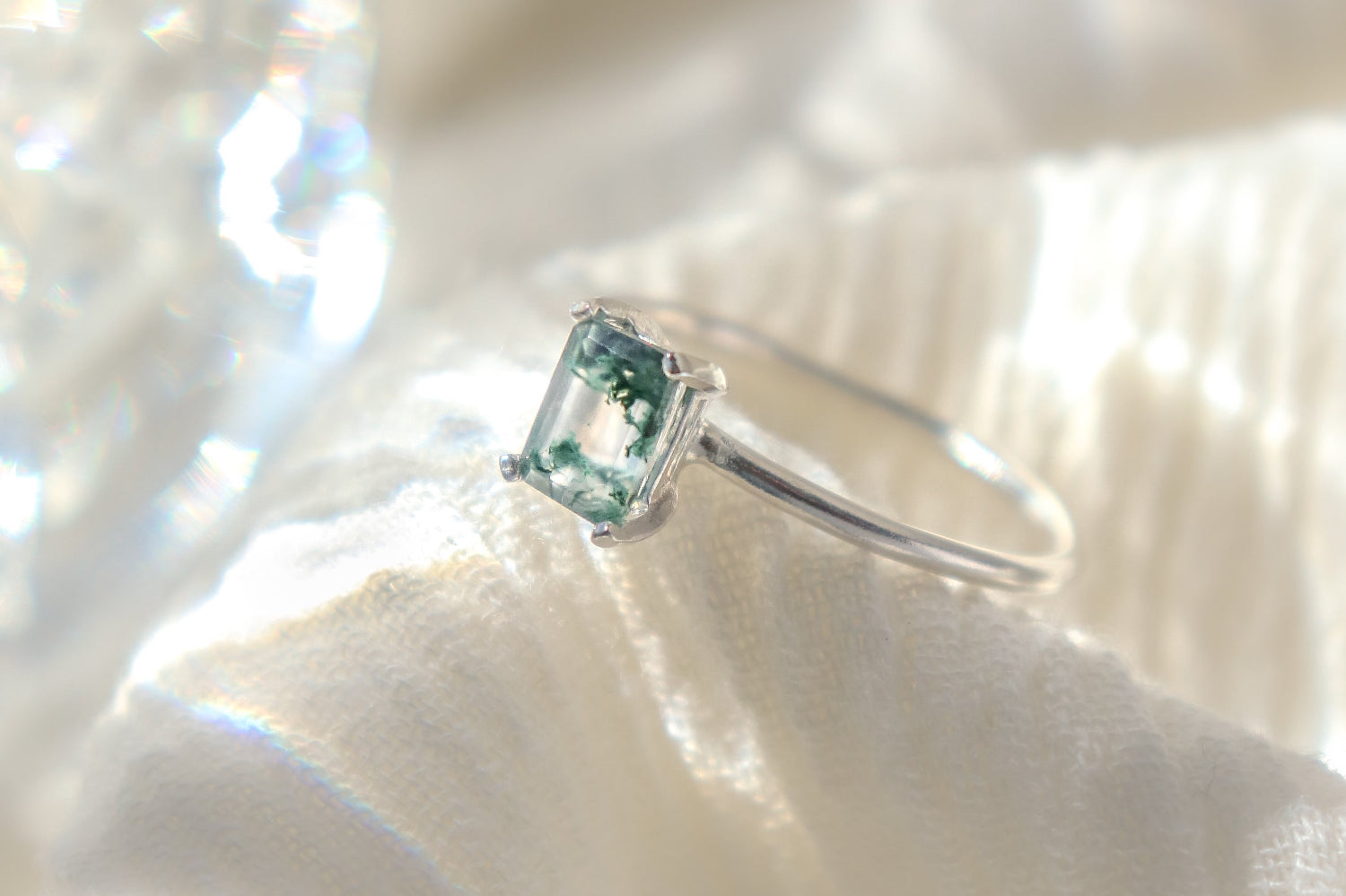 Rectangle shaped natural moss agate gemstone ring in a solid sterling silver setting with a smooth, round band. The ring is poised on a soft fabric background with moody lighting.