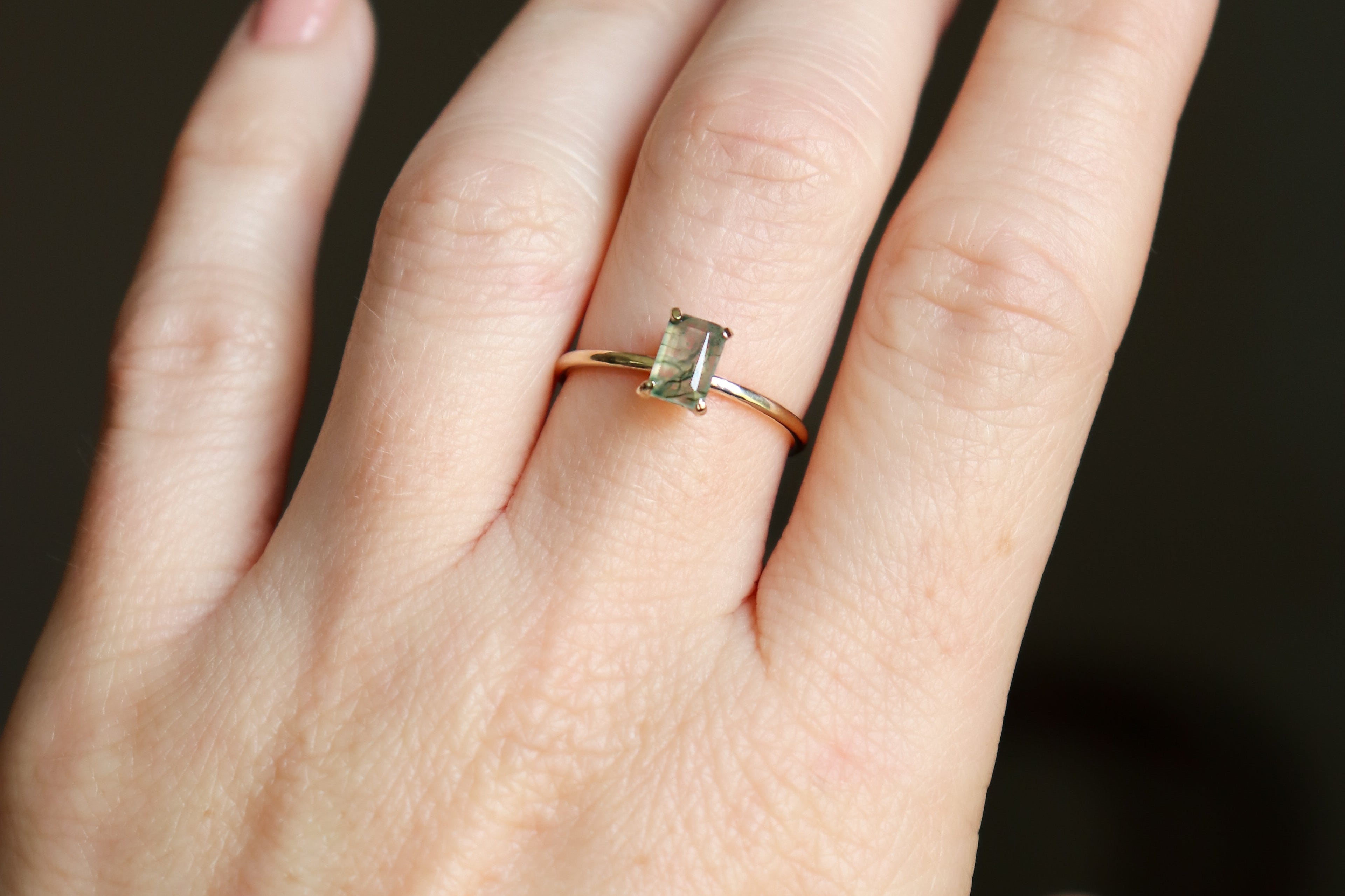 Solid gold moss agate ring on a woman's hand shown close up. The moss agate is a soft, ethereal, mossy fern green color with muted light filtering through the 4x6 mm emerald cut rectangle gemstone.