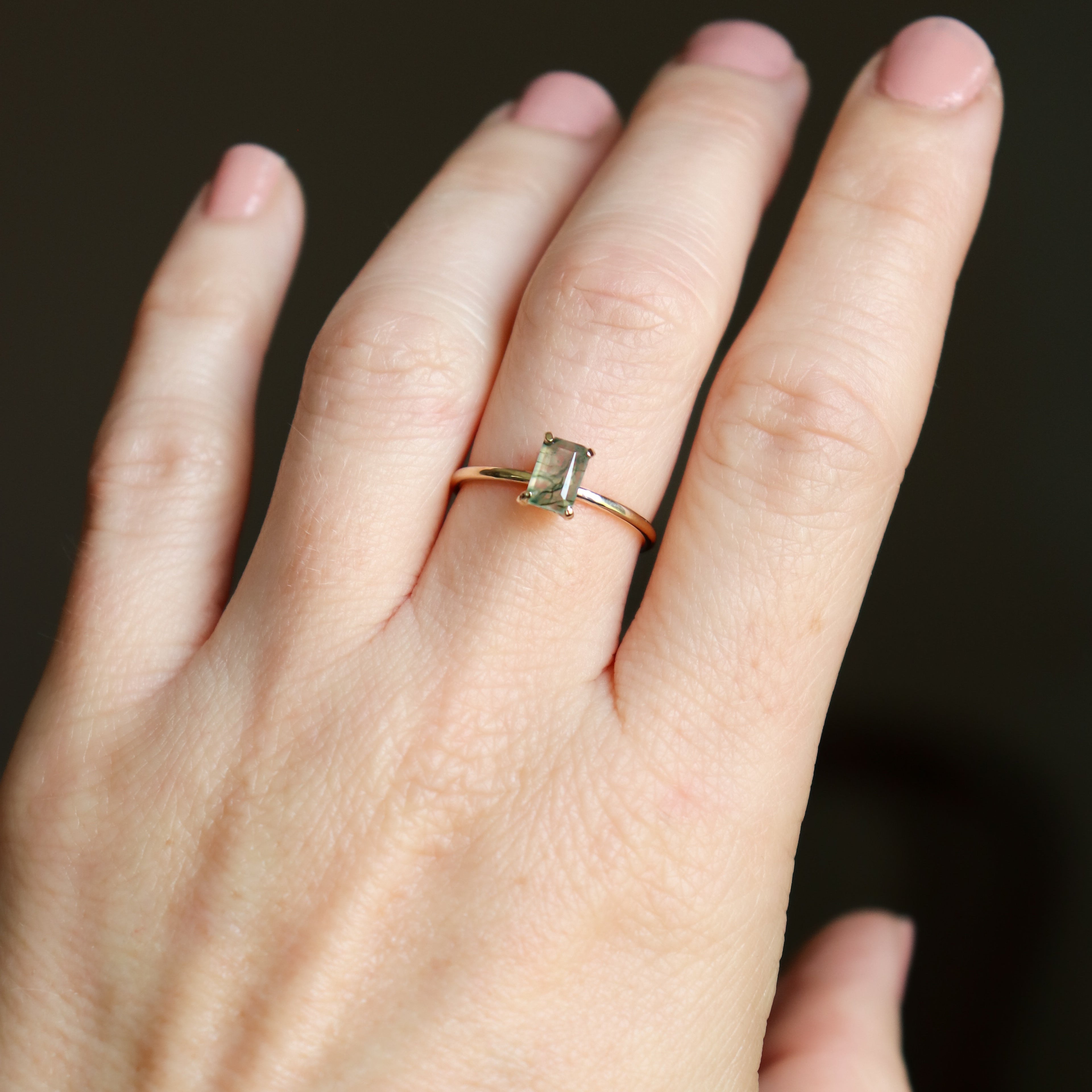Solid gold moss agate ring on a woman's hand shown close up. The moss agate is a soft, ethereal, mossy fern green color with muted light filtering through the 4x6 mm emerald cut rectangle gemstone.